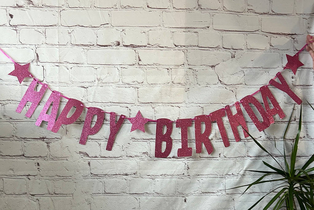 Happy Birthday banner with pink and red letters against a white brick wall.